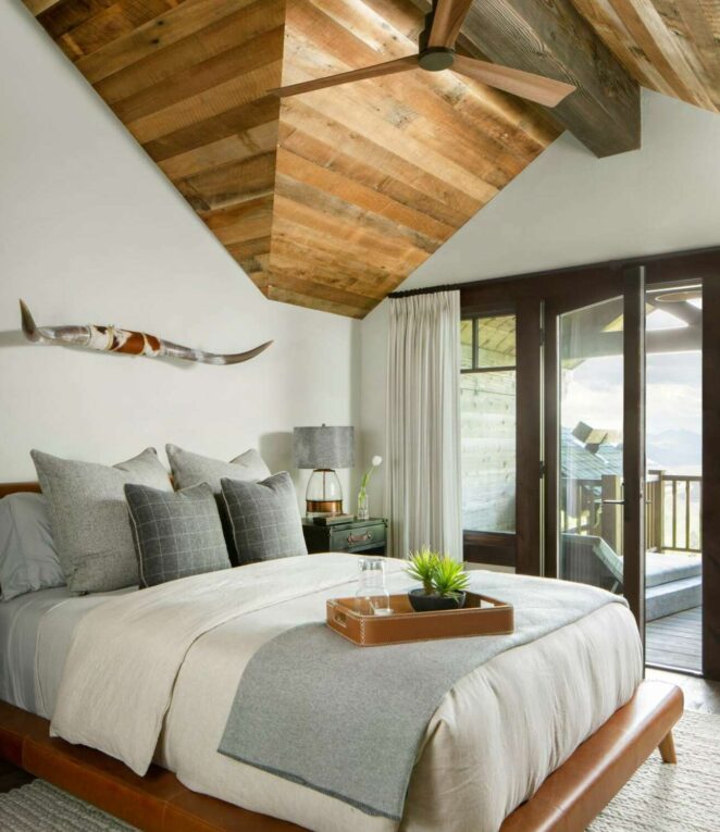 Image of primary bedroom with aged wood angled ceiling detail, longhorn steer horns, sliding door to balcony, and neutral colored linens on the bed; design by rebaL design, photography by Kimberly Gavin